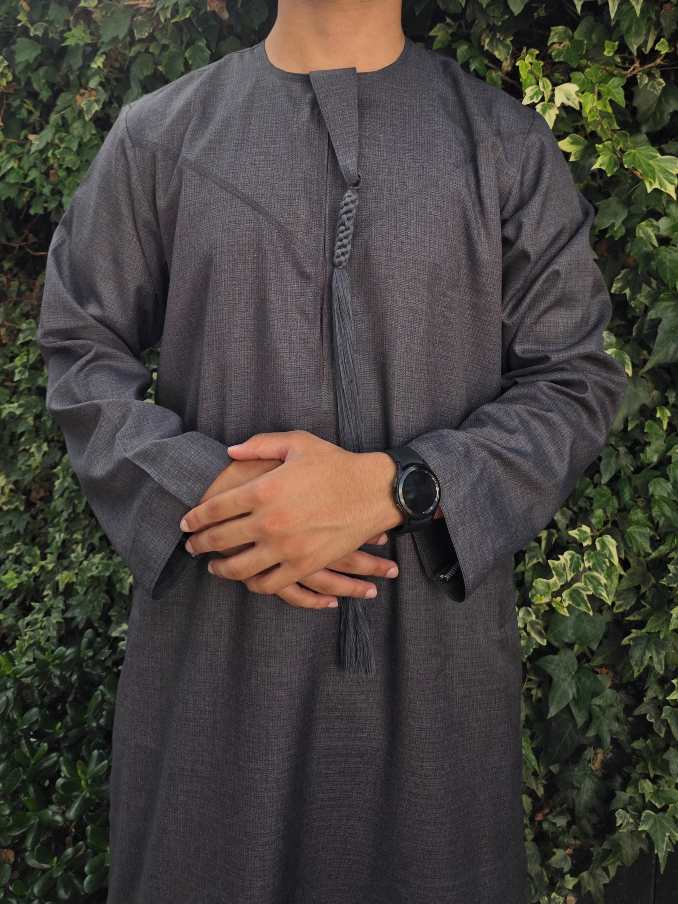 A person wearing a dark charcoal black Emirati thobe with a classic round collar and a sleek zip, featuring a traditional Emirati embroidery and a removable tassel.