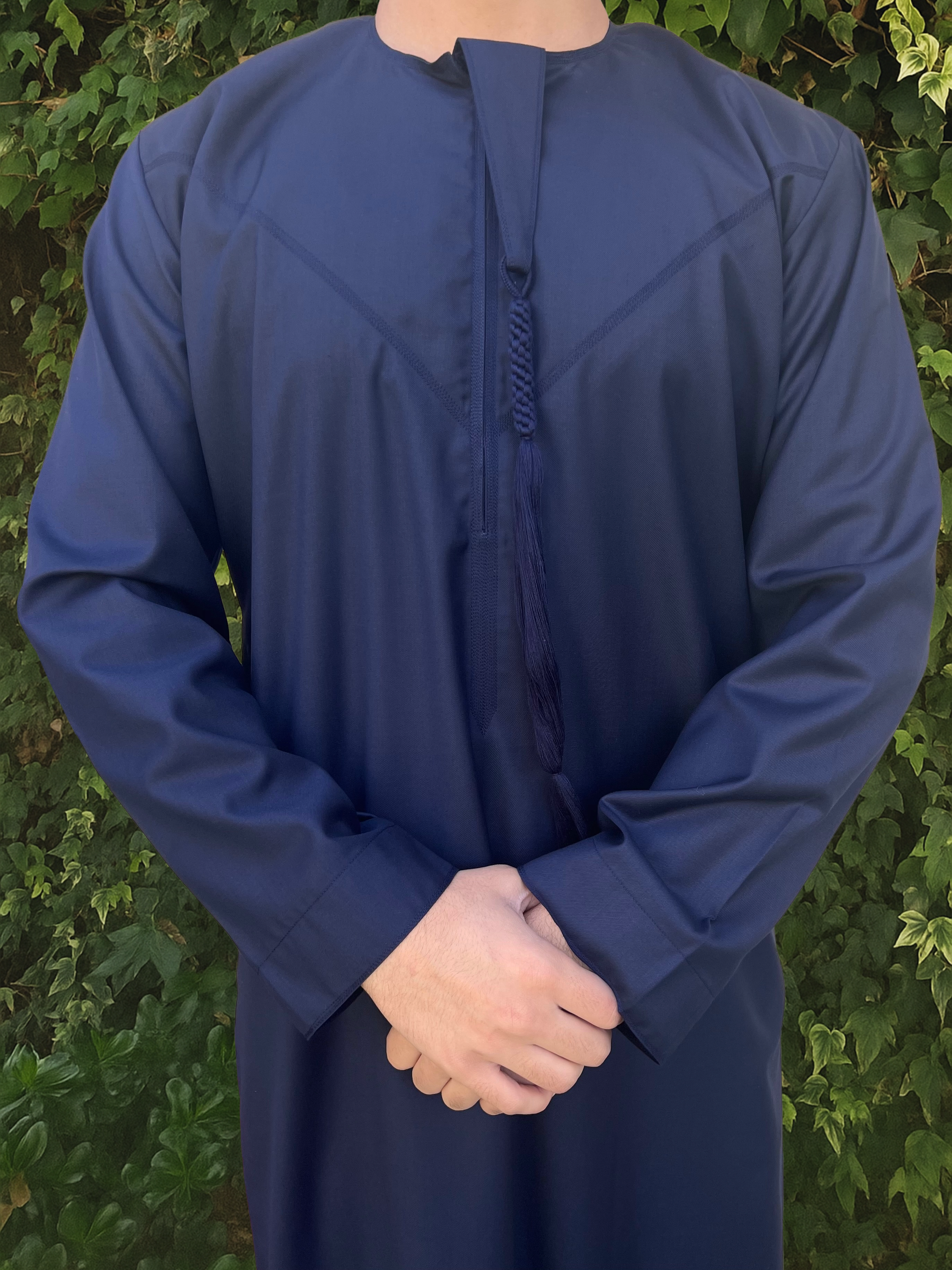 Person wearing a blue long-sleeve thobe against a green leafy background