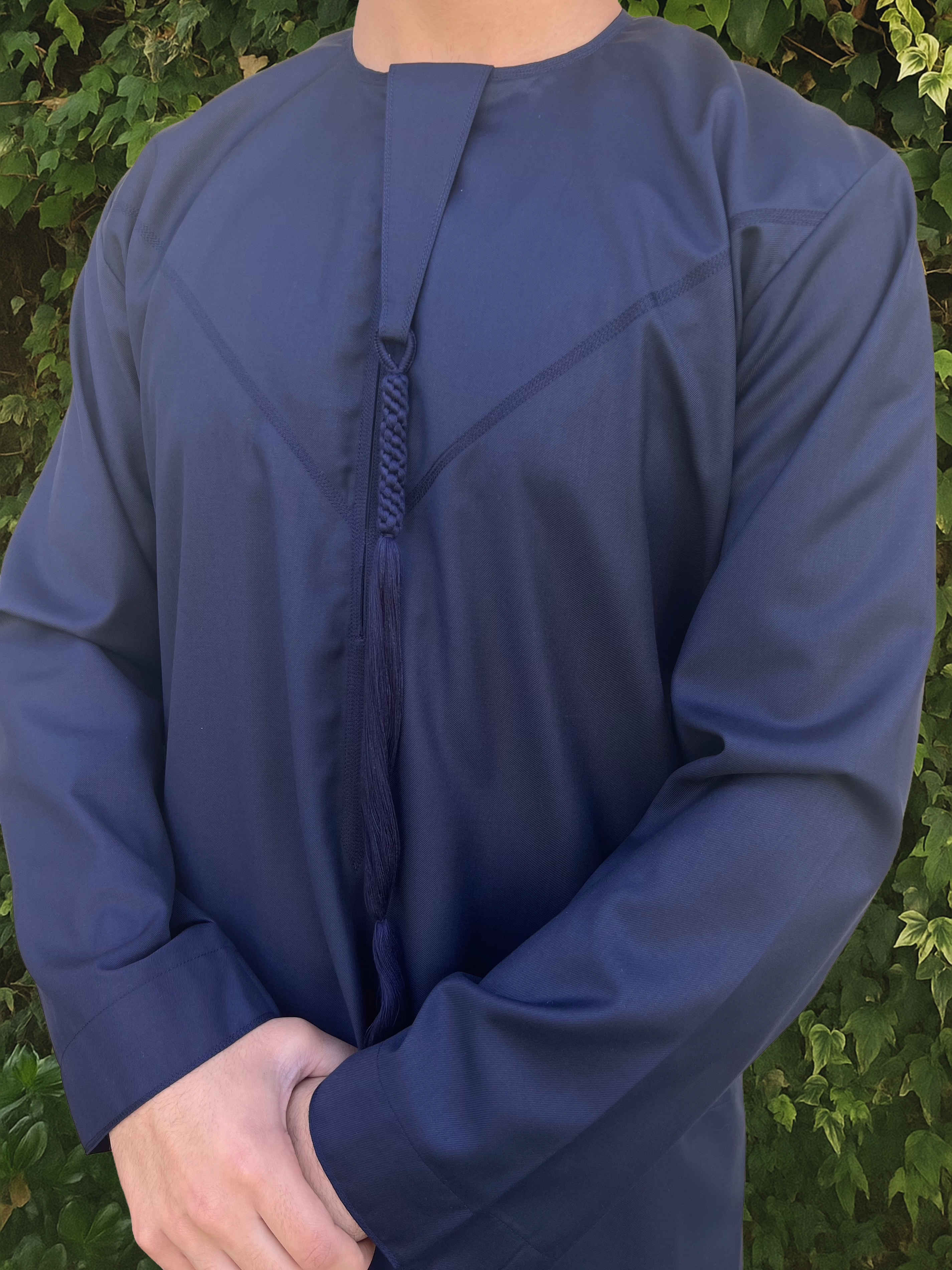 Person wearing a blue long-sleeve emirati thobe with a decorative blue detail against a green leafy background