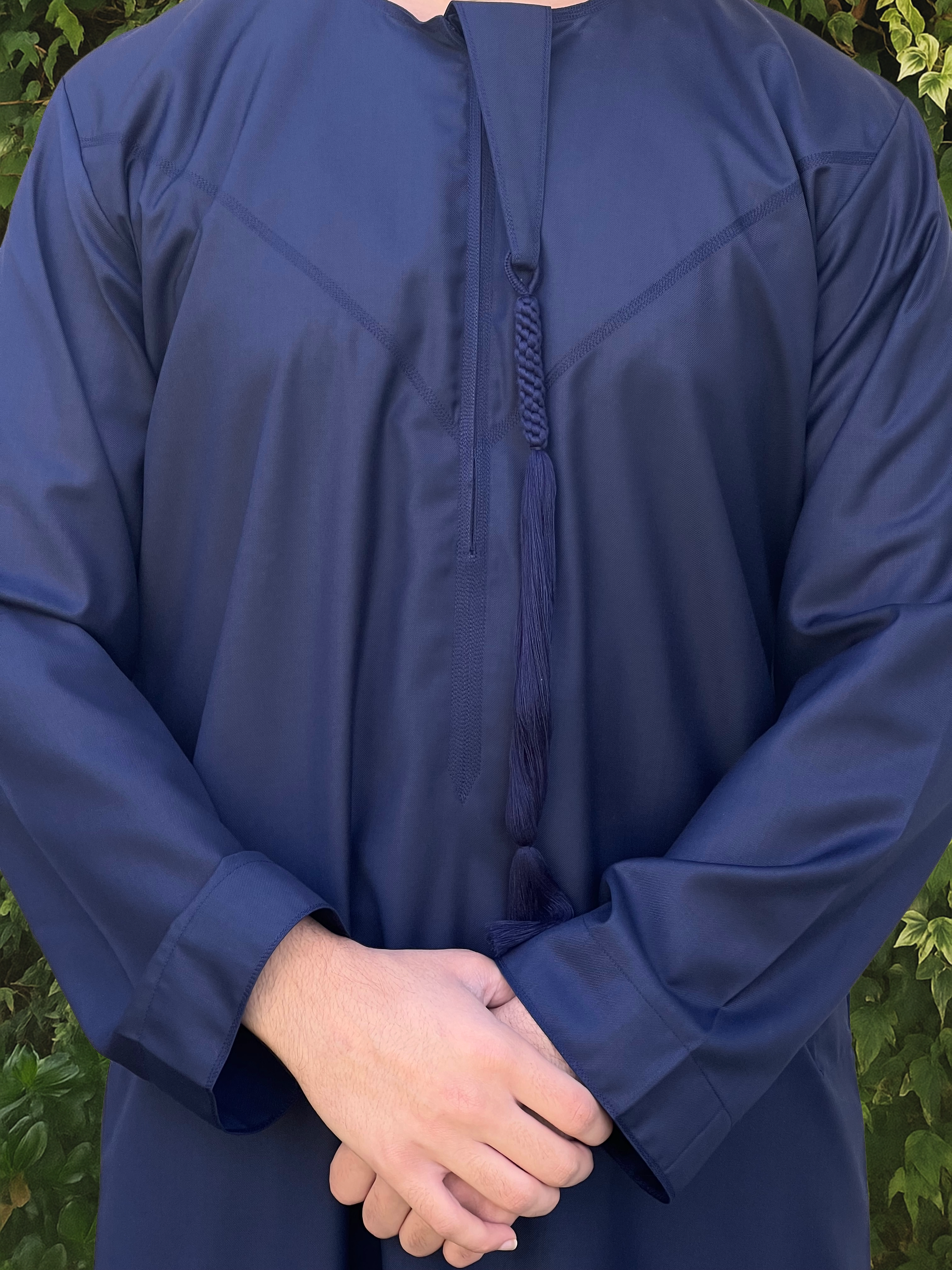 Person wearing a dark blue long-sleeve emirati thobe against a green leafy background