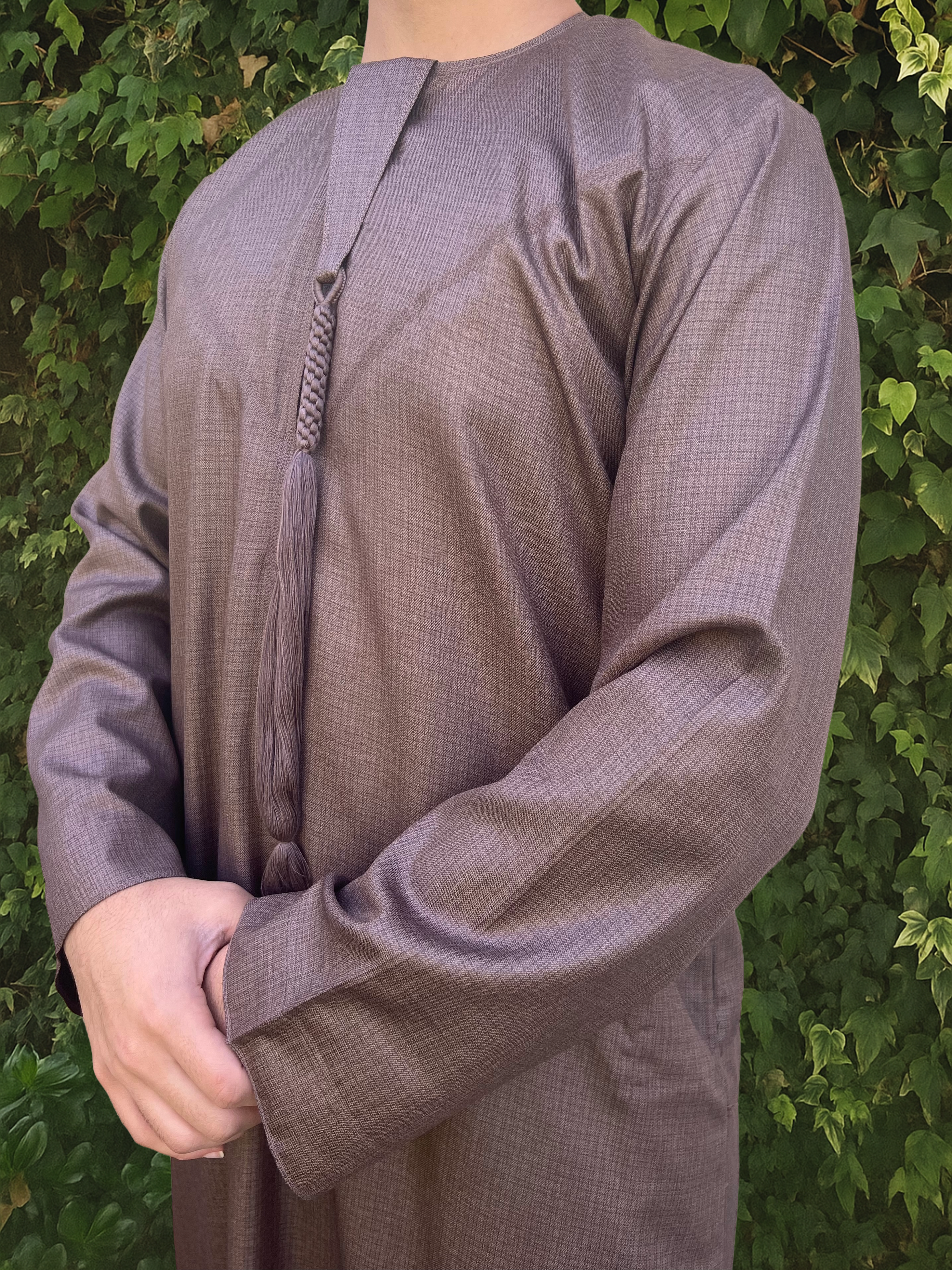 Person wearing a long-sleeve brown Emirati thobe with a tassel against a green leafy background